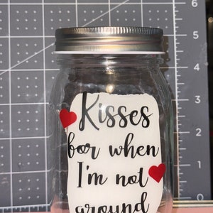 May include: A clear glass jar with a silver lid. The jar has a white label with the words "Kisses for when I'm not around" in black script, and two red heart stickers.