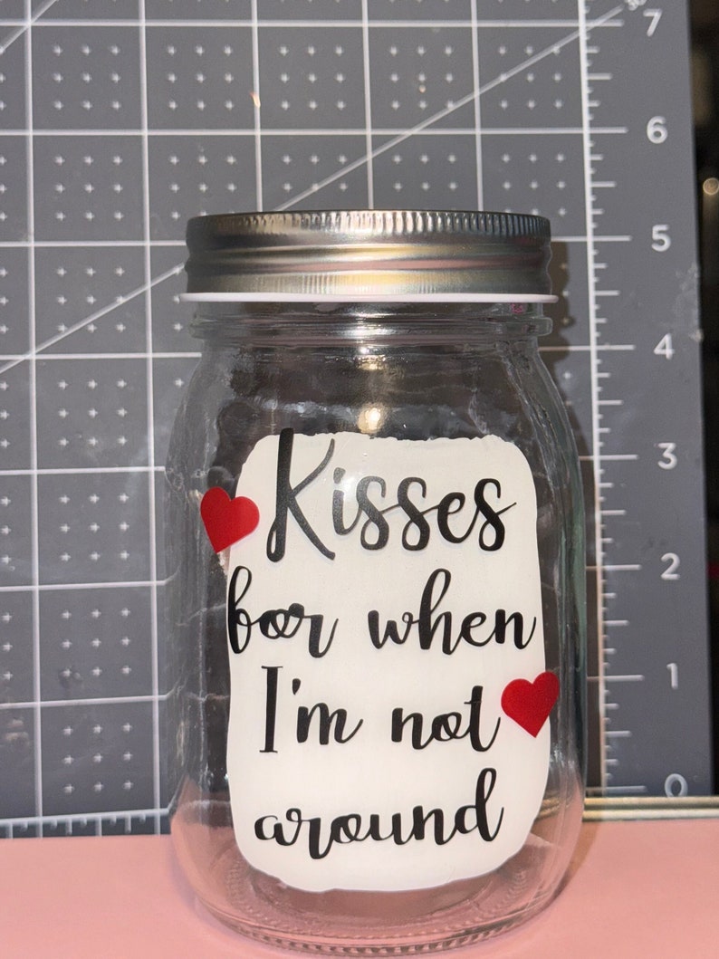 May include: A clear glass jar with a silver lid. The jar has a white label with the words "Kisses for when I'm not around" in black script, and two red heart stickers.