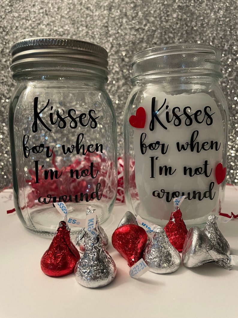 May include: Two clear glass mason jars with silver lids, each inscribed with "Kisses for when I'm not around" in black script. Red heart accents and foil-wrapped chocolate kisses are scattered around the jars, creating a romantic theme.