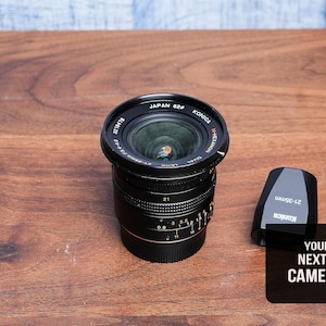 May include: A black Konica camera lens with the text "Japan 62#" and "Konica M-Hexanon Dual" is on a wooden surface. A black lens cap and a black sign that says "YOUR NEXT CAMERA" are also present.