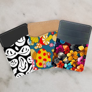May include: Three different card holders with unique designs. The first card holder has a black background with white smiley faces. The second card holder has a blue background with colorful mushrooms and flowers. The third card holder has a dark gray background with colorful flowers.