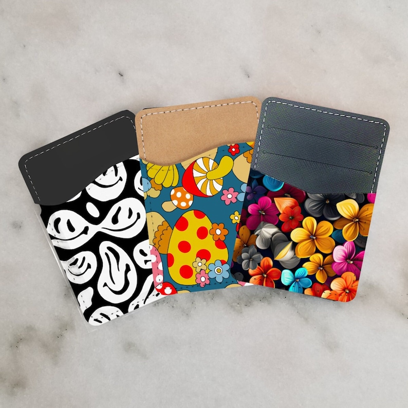 May include: Three different card holders with unique designs. The first card holder has a black background with white smiley faces. The second card holder has a blue background with colorful mushrooms and flowers. The third card holder has a dark gray background with colorful flowers.