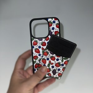 Peut inclure: Une coque de t&eacute;l&eacute;phone blanche avec une bordure noire et un motif de fraises rouges, de myrtilles et de m&ucirc;res. Un portefeuille en cuir noir est attach&eacute; &agrave; l'arri&egrave;re de la coque.