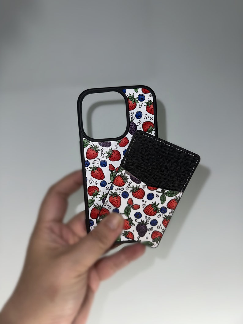 Peut inclure: Une coque de t&eacute;l&eacute;phone blanche avec une bordure noire et un motif de fraises rouges, de myrtilles et de m&ucirc;res. Un portefeuille en cuir noir est attach&eacute; &agrave; l'arri&egrave;re de la coque.