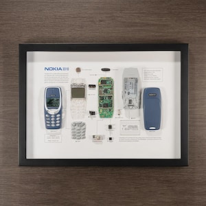 May include: A framed display showcasing the disassembled components of a Nokia 3310 phone. The components include the phone's blue casing, circuit board, battery, and keypad, all arranged on a white background with the text "NOKIA 3310".