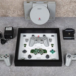 May include: A framed display featuring a disassembled PlayStation controller and components, with a gray console above. The display includes a controller, circuit board, and buttons. Additional controllers and accessories are arranged around the frame.