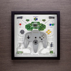 Può includere: Opera d'arte incorniciata con un controller Nintendo 64 smontato. Il controller grigio è al centro, circondato da componenti elettronici e pulsanti. La cornice è nera e l'opera include il logo Nintendo 64.