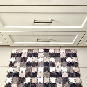 May include: A brown and white checkered kitchen rug with a stitched design. The rug is placed in front of a set of white kitchen cabinets with silver handles.