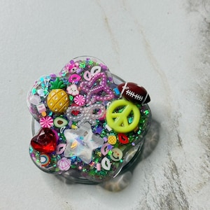 May include: A clear, heart-shaped phone grip filled with colorful glitter and miniature charms. The charms include a yellow pineapple, a red heart, a peace sign, a football, and various candies and stars. The grip is designed to attach to the back of a phone.