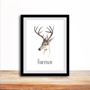 Op de afbeelding: Een ingelijst aquarel schilderij van een hert met geweien. Het hert kijkt naar links en het schilderij staat op een houten oppervlak. De naam "Harrison" staat onder het hert gedrukt.