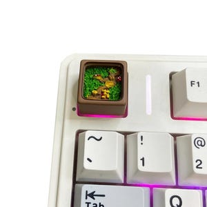 May include: A custom keyboard keycap with a miniature forest diorama, including green moss, mushrooms, and a red mushroom. The brown keycap is set on a white keyboard with pink underlighting. The keycap is a unique accessory.