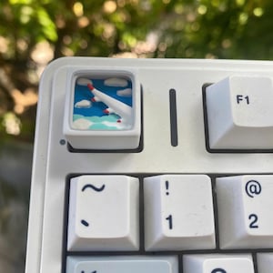 Könnte beinhalten: Weiße mechanische Tastatur mit einer individuellen Tastenkappe, die ein stilisiertes Flugzeug zeigt, das durch Wolken fliegt. Die Tastenkappe hat einen blauen Himmelshintergrund mit weißen Wolken und einem rotspitzigen Flugzeug. Andere Tasten sind weiß mit schwarzer Beschriftung.