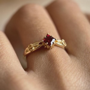 May include: A gold ring featuring a square, deep red gemstone as the focal point. The band has an intricate, openwork design with small accent stones. The ring is shown on a finger, highlighting the jewelry's details and craftsmanship.