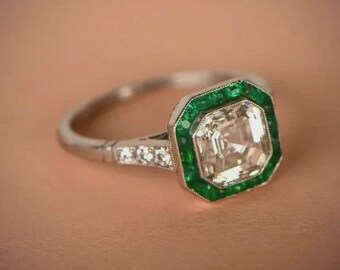 Könnte beinhalten: Ein silberner Ring mit einem großen, quadratischen, klaren Edelstein, umgeben von einem Rand aus grünen Edelsteinen. Der Ring hat ein detailliertes Band mit kleinen, klaren Edelsteinen. Der Ring ist vor einem neutralen Hintergrund platziert.