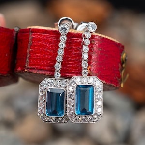 Pode incluir: Um par de brincos pendentes com uma gema de topázio azul cercada por um halo de diamantes. Os brincos são feitos de prata e têm um design de gota.