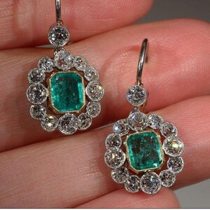 May include: A pair of emerald and diamond drop earrings. The earrings feature a square emerald gemstone surrounded by a halo of round diamonds. The earrings are suspended from a delicate silver chain.