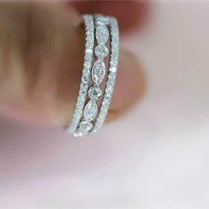 Puede incluir: Primer plano de un anillo plateado con tres bandas. Las bandas exteriores están incrustadas con pequeñas piedras redondas y transparentes. La banda central presenta piedras claras más grandes, de forma ovalada. El anillo se sostiene sobre un fondo rosa suave.