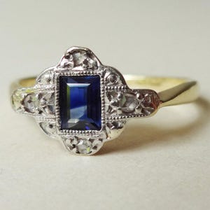 May include: A vintage-style ring with a rectangular, deep blue gemstone at the center. The gemstone is set in a decorative, silver-toned metal setting, accented with smaller, clear stones. The band is gold-toned, creating a classic, elegant look.