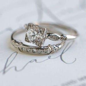 May include: Two silver rings, one with a square-cut diamond centre stone and a smaller band with embedded diamonds. The rings are set on a white surface with cursive writing, showcasing a classic and elegant jewellery design.