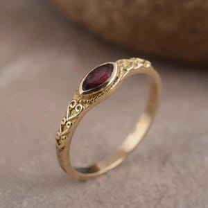 May include: A gold ring with a maroon oval-shaped gemstone. The ring has an ornate, filigree design along the band. The gemstone is set in a bezel setting. The ring is on a neutral background.