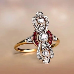 May include: A gold ring with a central design featuring a large, round, clear gemstone surrounded by smaller clear gemstones and red gemstones. The ring has an ornate, vintage style with a detailed setting.