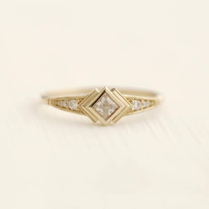 Puede incluir: Un anillo de oro con un diamante de forma cuadrada en el centro y diamantes más pequeños engastados a lo largo de la banda.