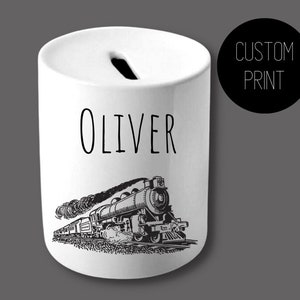 Puede incluir: Hucha de cerámica blanca con una ranura en la parte superior. El nombre "Oliver" está impreso en negro, encima de una ilustración en blanco y negro de un tren de vapor. Un círculo negro con "CUSTOM PRINT" está a la derecha.