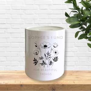 May include: White ceramic money bank with a floral design and the text "SOPHIE'S FUND FOR BEAUTIFUL THINGS". The bank has a slot at the top for inserting money. The floral design is black and white.