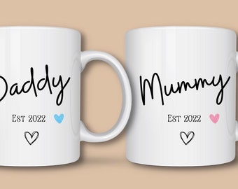 May include: Two white ceramic mugs with black text that reads "Daddy" and "Mummy". Each mug has "EST 2022" below the names, a heart, and a blue or pink heart icon. The mugs are for a new parent.