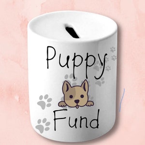 Peut inclure: Tirelire en céramique blanche avec une fente sur le dessus. Elle affiche l'inscription "Puppy Fund" en noir, un dessin de chiot et des empreintes de pattes.