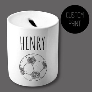 May include: White ceramic money bank with a soccer ball design. The name "HENRY" is printed in black above the soccer ball. A black circle with the words "CUSTOM PRINT" is on the right side.