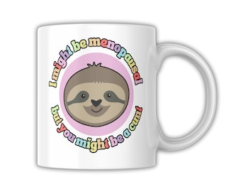 Puede incluir: Taza de cerámica blanca con la cara de un perezoso de dibujos animados dentro de un círculo rosa, con el texto "I might be menopausal but you might be a cunt" en arcoíris. La taza tiene un asa blanca.