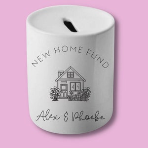 Puede incluir: Hucha de cerámica blanca con una ranura en la parte superior. La hucha presenta el texto "NEW HOME FUND" encima de un dibujo lineal de una casa con flores. Debajo de la casa está el texto "Alex & Phoebe".