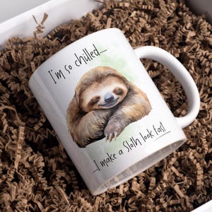 May include: White ceramic mug with a watercolour illustration of a sleeping sloth. The mug has the text "I'm so chilled... I make a Sloth look fast". The mug is inside a white box with brown crinkle paper.