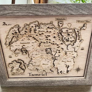 May include: A wooden map of the fictional continent of Tamriel from the Elder Scrolls video game series. The map is framed in a light brown wood frame and features the names of the various provinces, including Skyrim, Cyrodiil, Morrowind, and Black Marsh.