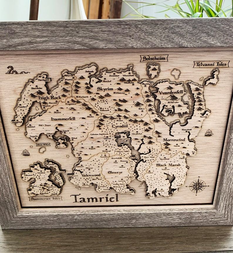 May include: A wooden map of the fictional continent of Tamriel from the Elder Scrolls video game series. The map is framed in a light brown wood frame and features the names of the various provinces, including Skyrim, Cyrodiil, Morrowind, and Black Marsh.