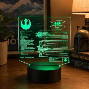 Puede incluir: Una lámpara de pantalla acrílica LED de color verde brillante con un esquema técnico de un X-Wing Starfighter. El panel transparente muestra diagramas detallados, etiquetas y el símbolo de la Alianza Rebelde, montado sobre una base negra circular.