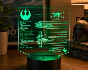 Könnte beinhalten: Eine leuchtend grüne LED-Acryl-Displaylampe mit einem technischen Schaltplan eines X-Wing Starfighters. Die transparente Platte zeigt detaillierte Diagramme, Beschriftungen und das Symbol der Rebellenallianz auf einem runden schwarzen Sockel.