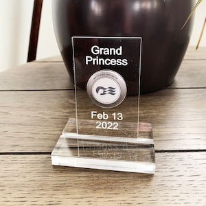 May include: A clear acrylic keepsake with the words "Grand Princess" in white. A circular emblem with a ship logo and the text "Anthony Hoiseth" and "Discovery Princess - Feb 25, 2023" is below the text. The date "Feb 13 2022" is printed below the emblem.