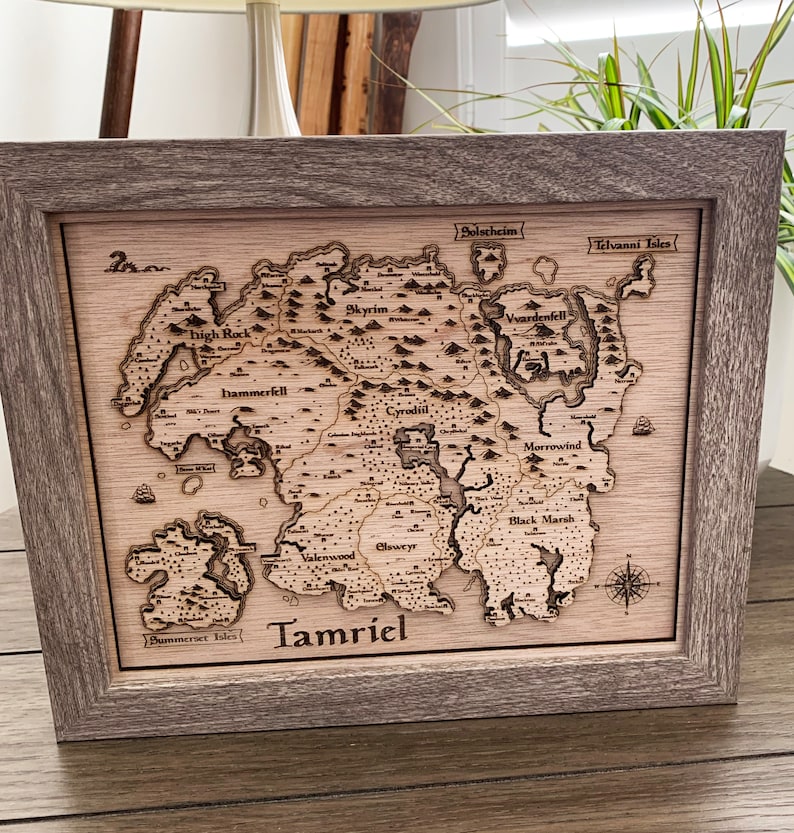 May include: A wooden map of Tamriel, the fictional continent from the Elder Scrolls video game series. The map is framed in a light brown wood frame and features the names of the various provinces, including Skyrim, Cyrodiil, Morrowind, and Hammerfell.