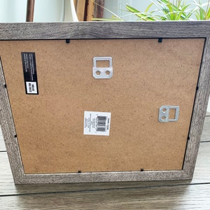 May include: A gray wood picture frame with a brown cardboard backing. The frame has two metal hanging hooks on the back. The frame has a label that reads "FRAME SIZE 12X16" and "SIZE 12X16".