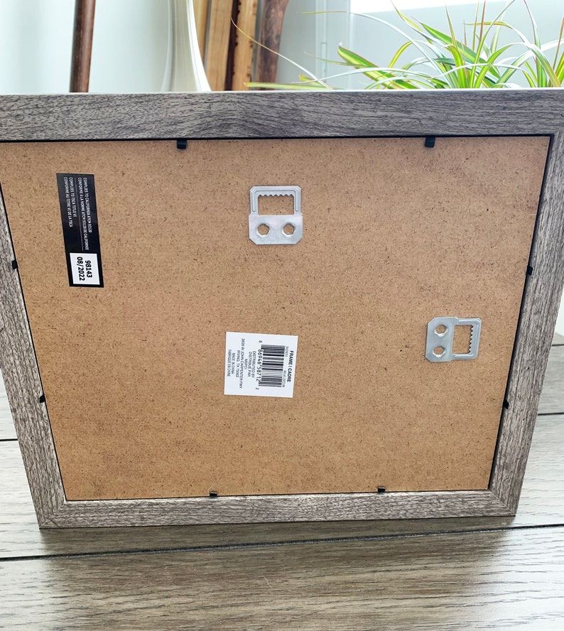 May include: A gray wood picture frame with a brown cardboard backing. The frame has two metal hanging hooks on the back. The frame has a label that reads "FRAME SIZE 12X16" and "SIZE 12X16".