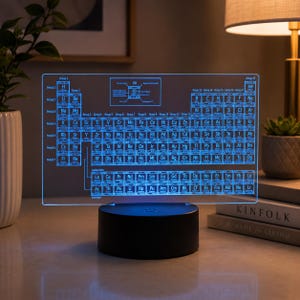 May include: A rectangular acrylic display featuring an illuminated blue periodic table of elements. The clear panel is mounted on a solid black cylindrical base with a power button. The table includes chemical symbols, atomic numbers, and a key.
