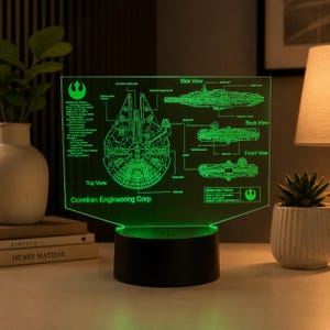 Könnte beinhalten: Eine grüne LED-Acryllampe mit einem technischen Bauplan des Millennium Falcon. Das Display zeigt Ansichten von oben, von der Seite, von hinten und von vorne mit Beschriftungen wie Corellian Engineering Corp und verschiedenen Schiffskomponenten auf einem schwarzen Sockel.