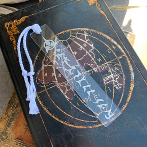 May include: A clear acrylic bookmark with a white design of a stylized tree and the word "STEELE" on it. The bookmark is attached to a white string.