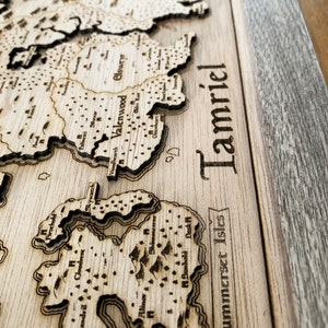 May include: A wooden map of Tamriel, the continent from the Elder Scrolls video game series. The map is laser-cut and features the names of various regions and cities, including Valenwood, Elsweyr, and Summerset Isles.