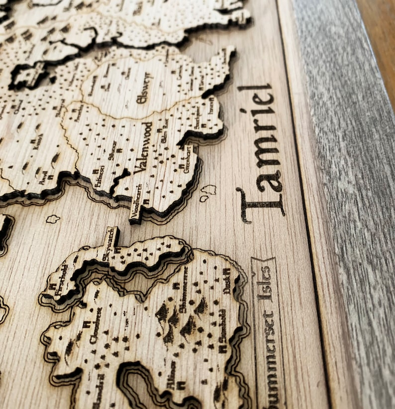 May include: A wooden map of Tamriel, the continent from the Elder Scrolls video game series. The map is laser-cut and features the names of various regions and cities, including Valenwood, Elsweyr, and Summerset Isles.