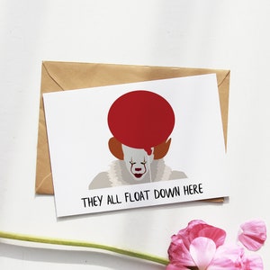 Könnte beinhalten: Eine weiße Karte mit einem roten Ballon und einem Clown-Gesicht mit dem Text "They all float down here". Die Karte liegt auf einem braunen Umschlag, um den herum rosafarbene Blütenblätter verstreut sind.