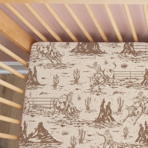 May include: A beige and brown crib sheet with a repeating pattern of cowboys riding bulls. The pattern is a vintage style illustration.
