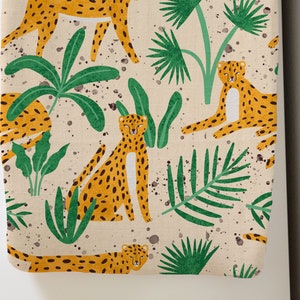 May include: A beige fabric with a repeating pattern of yellow cheetahs with black spots and green tropical plants. The design includes palm fronds and other foliage, with brown speckles scattered across the background.